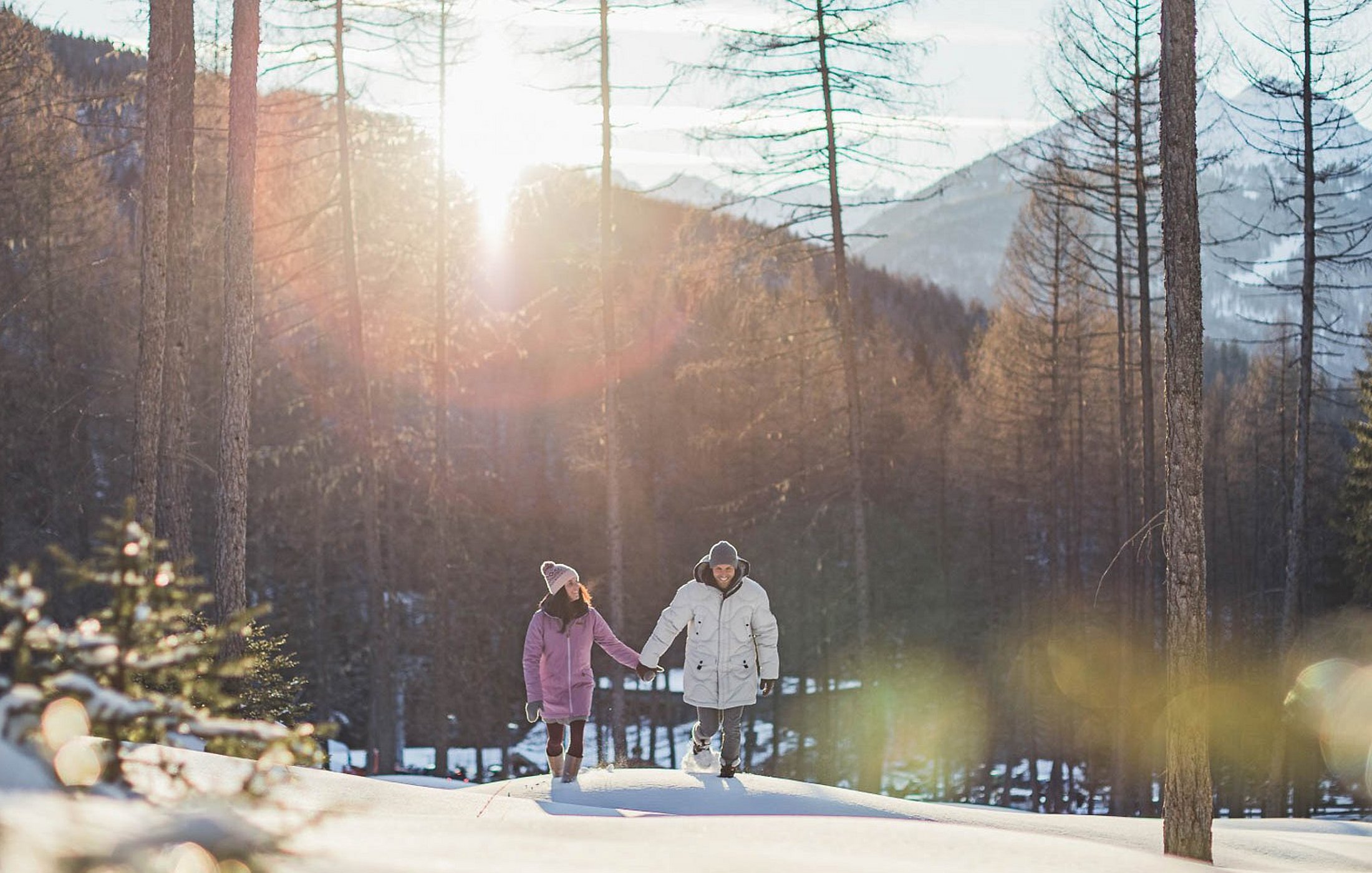 Paar beim Winterspaziergang im Wald mit Sonne im Hintergrund und verschneiten Bäumen